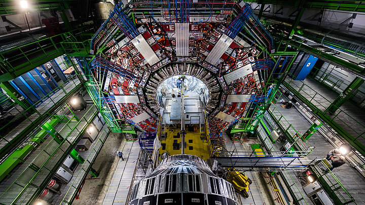 Zdjęcie ogromnej hali, w której w centralnym miejscu znajduje się cylindryczny element aparatury wielkiego zderzacza hadronów. Hala wypełniona jest wielopoziomowymi rusztowaniami zapewniającymi pracownikom dostep do najwyzszych poziomów aparatury. 
