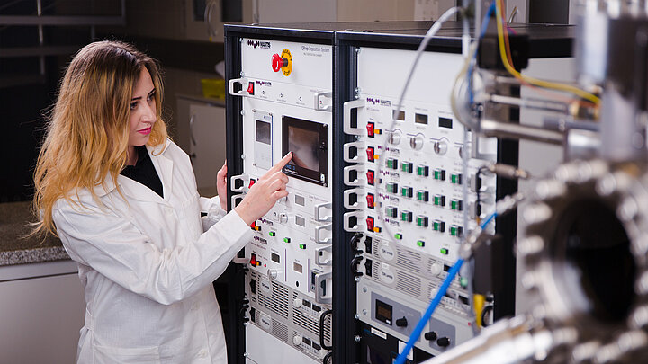 Zdjęcie przedstawia kobietę ubraną w biały fartuch, stojącą obok aparatury badawczej. Wskazuje palcem na dane na wyświetlaczu.