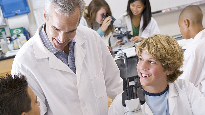 Sala laboratoryjna. Nauczyciel tłumaczy coś uczniom pracującym z mikroskopami. 